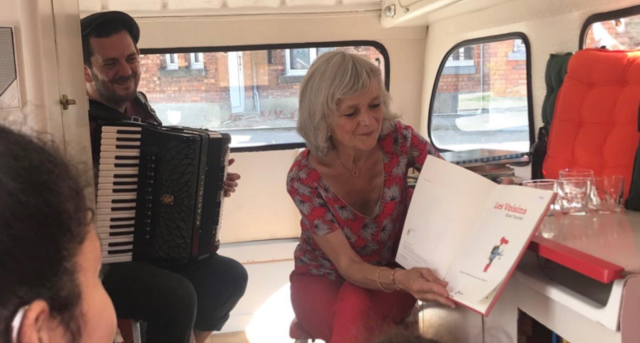 Une dame lit, et un monsieur joue de l'accordéon dans une caravane